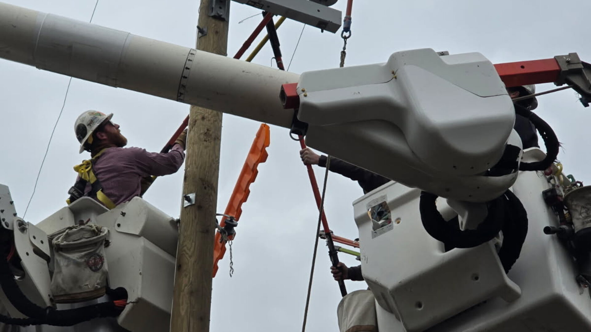 Two linemen in bucket trucks using insulated hot sticks on a wood pole with orange protective line covers